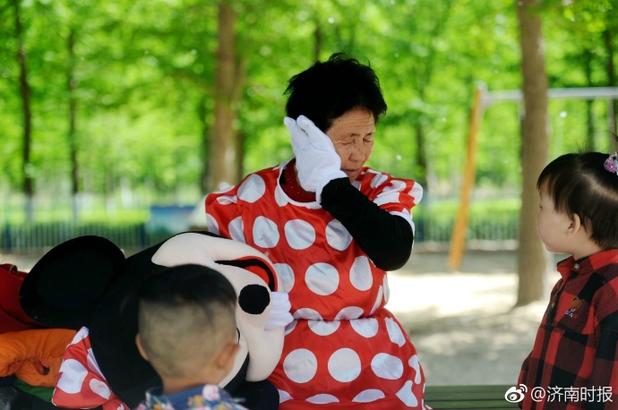 18日,在济南张庄路附近的森林公园里,一位老人身穿米老鼠的演出服,戴上厚重的面具,邀请过往的游客与其合影,同时向游客讲述家庭遭遇,为病床上的儿媳妇募集治疗费。